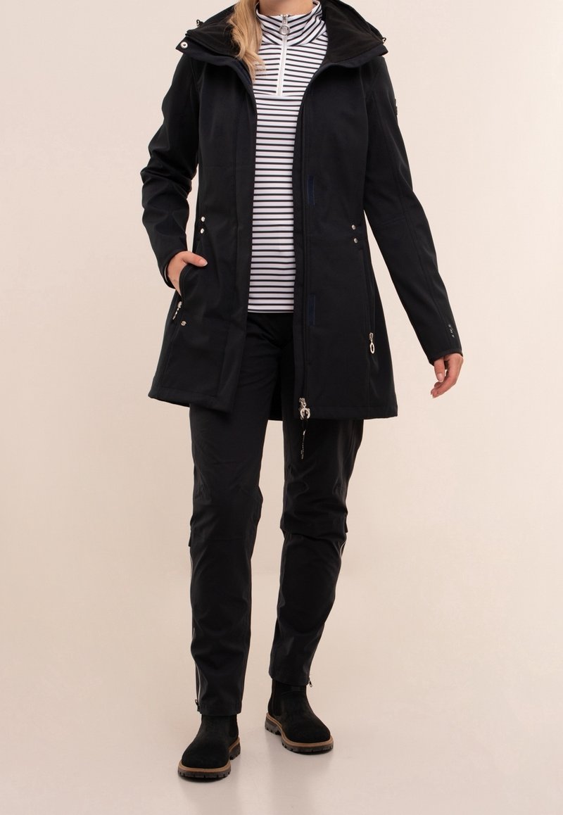 Vrouw in een lange zwarte jas, zwarte broek, zwarte laarzen en een wit-zwarte gestreepte zip-up top, staande op een lichte achtergrond.