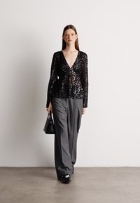 Μαύρο διάφανο τοπ με μακριά μανίκια και στροβιλιζόμενο σχέδιο, V λαιμόκοψη, συνδυασμένο με γκρι ριγέ παντελόνι φαρδύ και μια μικρή μαύρη τσάντα.