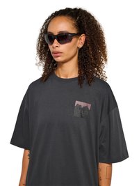 Kobieta z kręconymi włosami, nosząca czarne okulary przeciwsłoneczne oraz luźną ciemnoszarą koszulkę z małym nadrukiem na piersiach.