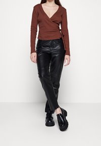 Mujer vistiendo una blusa envolvente de color óxido, pantalones de cuero negros y mocasines negros gruesos, de pie contra un fondo blanco liso.