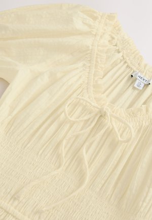 Vaaleankeltainen yläosa, joka on valmistettu teksturoidusta kankaasta. Siinä on röyhelöreunainen kaula-aukko, smokattu koriste ja edessä säädettävä nauha.