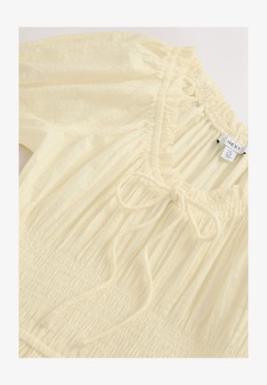 Vaaleankeltainen yläosa, joka on valmistettu teksturoidusta kankaasta. Siinä on röyhelöreunainen kaula-aukko, smokattu koriste ja edessä säädettävä nauha.