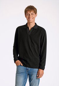 Svart långärmad pikétröja med texturerad ribbad yta och krage, kombinerad med blå jeans. Modellen står med ena handen i fickan.