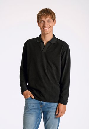 Polo shirt nero a maniche lunghe con coste testurizzate e colletto, abbinato a jeans blu. Il modello è in piedi con una mano in tasca.