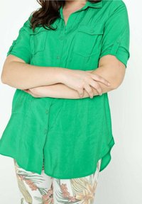 Donna con lunghi capelli scuri che indossa una camicia verde brillante con bottoni e pantaloni a fantasia floreale, le braccia incrociate in vita.