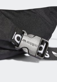 Närbild av svart tygrem med silverfärgad plastspänne graverat med "adidas" och svart rem med vit adidas-logotyp och text.