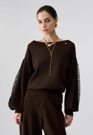 Donna con capelli tirati indietro indossa un abito marrone scuro con maniche a palloncino in pizzo trasparente e collane d'oro a strati e orecchini a cerchio.
