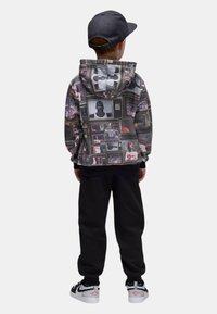 La felpa con cappuccio presenta un collage di schermi TV vintage in scala di grigi con accenti colorati, abbinata a pantaloni della tuta neri e sneakers nere.