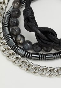 Tre lagdelte armbånd bestående af sølvkædelinks, sort læder og mørke perler med en glat tekstur. Bemærkelsesværdige blandede materialer og design.