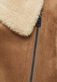 Veste en daim marron doux avec col en peau lainée crème et fermeture éclair en métal noir partiellement zippée sur le devant.