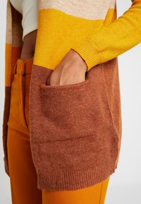 Persoon draagt een geblokte cardigan in geel, beige en roestkleur met hand in een grote voorzak, gecombineerd met mosterdgele broek.