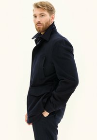 Laivansinisestä villasekoitekankaasta valmistettu takki, jossa on korkea kaulus, suuri etutasku ja muotoiltu istuvuus. Kankaassa on sileä rakenne ja hienovarainen kiilto.