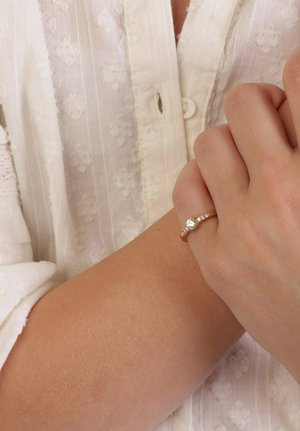 Gouden ring met een enkele centrale steen en kleinere accentstenen aan beide zijkanten. De hand rust op een stof met lichte textuur.