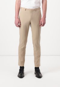 Pantaloni beige su misura con vestibilità slim, caratterizzati da una parte frontale liscia, passanti per la cintura e una texture morbida. Abbinati a scarpe nere e calze.