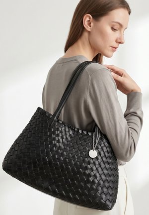 Jonge vrouw met lang bruin haar draagt een grijze trui en houdt een grote zwarte gevlochten leren schoudertas met een zilveren ronde label vast.
