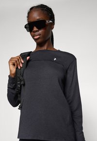 Young woman wearing large black sunglasses and a long-sleeve dark shirt, holding a black backpack strap over her shoulder.