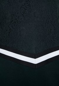 Un vestito verde scuro presenta una parte superiore in pizzo e una parte inferiore nera testurizzata. Ha un collo a V con bordi a strisce bianche e nere.