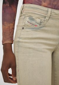 Jeans en denim beige avec des détails de couture rouge, dotés d'une étiquette de logo sur la poche avant et d'une coupe slim. Texture douce et finition lisse.