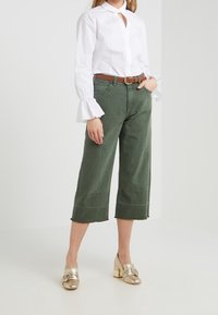 Nainen, joka on pukeutunut valkoiseen pitkähihhaiseen paitaan, jossa on levenevät hihansuut, vihreisiin leveisiin capris-housuihin, ruskeaan vyöhön ja kultaisiin korkokenkiin, seisoo valkoista taustaa vasten.