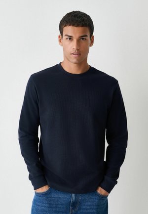 Navy blue long-sleeve T-shirt in a textured fabric, featuring a round neckline and cuffed sleeves, paired with blue denim jeans.