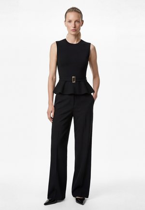 Femme debout, une main dans la poche, portant un haut peplum sans manches noir avec ceinture et un pantalon large noir, fond blanc uni.