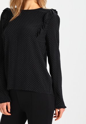Zwarte blouse met lange mouwen en kleine witte polka dots, met ruches op schouders en manchetten, gedragen met een zwarte broek.