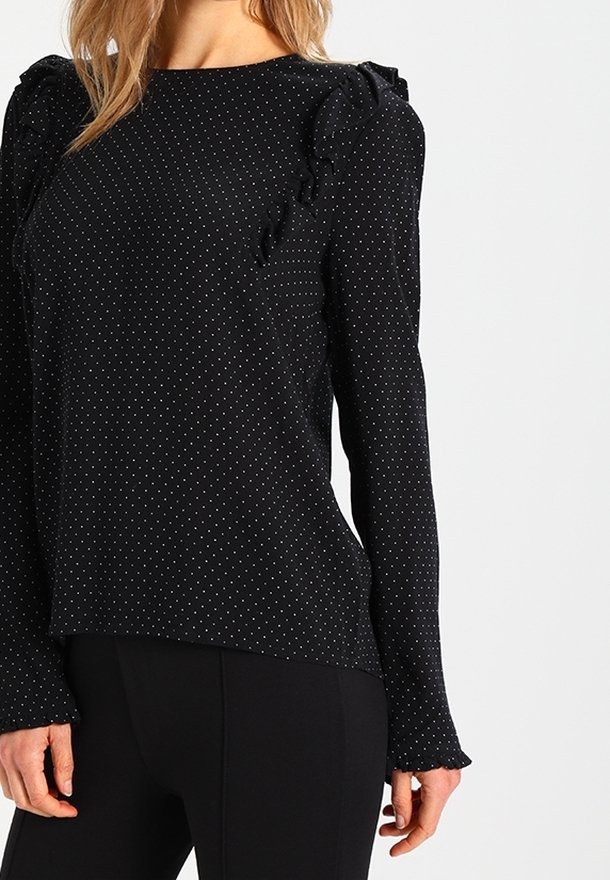 Blouse noire à manches longues à petits pois blancs avec des détails à volants sur les épaules et les poignets, portée avec un pantalon noir.