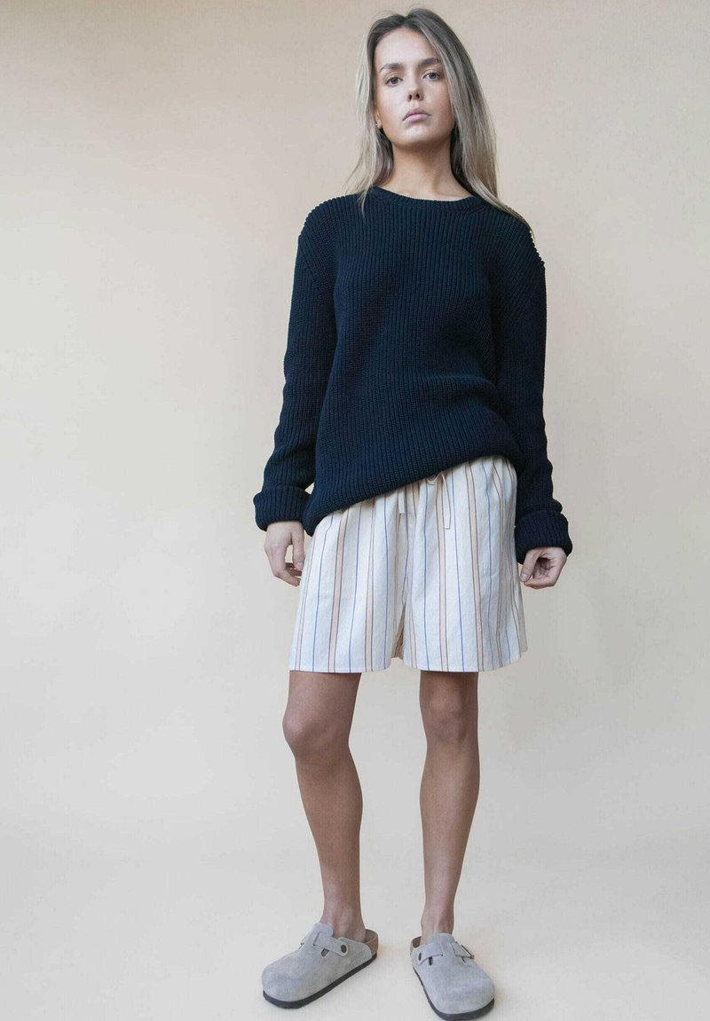 Marineblauwe ribbelgebreide trui, crèmekleurige gestreepte shorts met veelkleurige lijnen en grijze instappers. Ontspannen pasvorm met textuurlijke materialen.