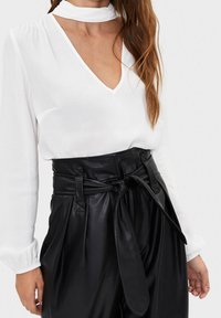 Vit blus med djup V-ringning och långa ärmar, kombinerad med högmidjade svarta byxor i fusk läder med en knuten bälte.