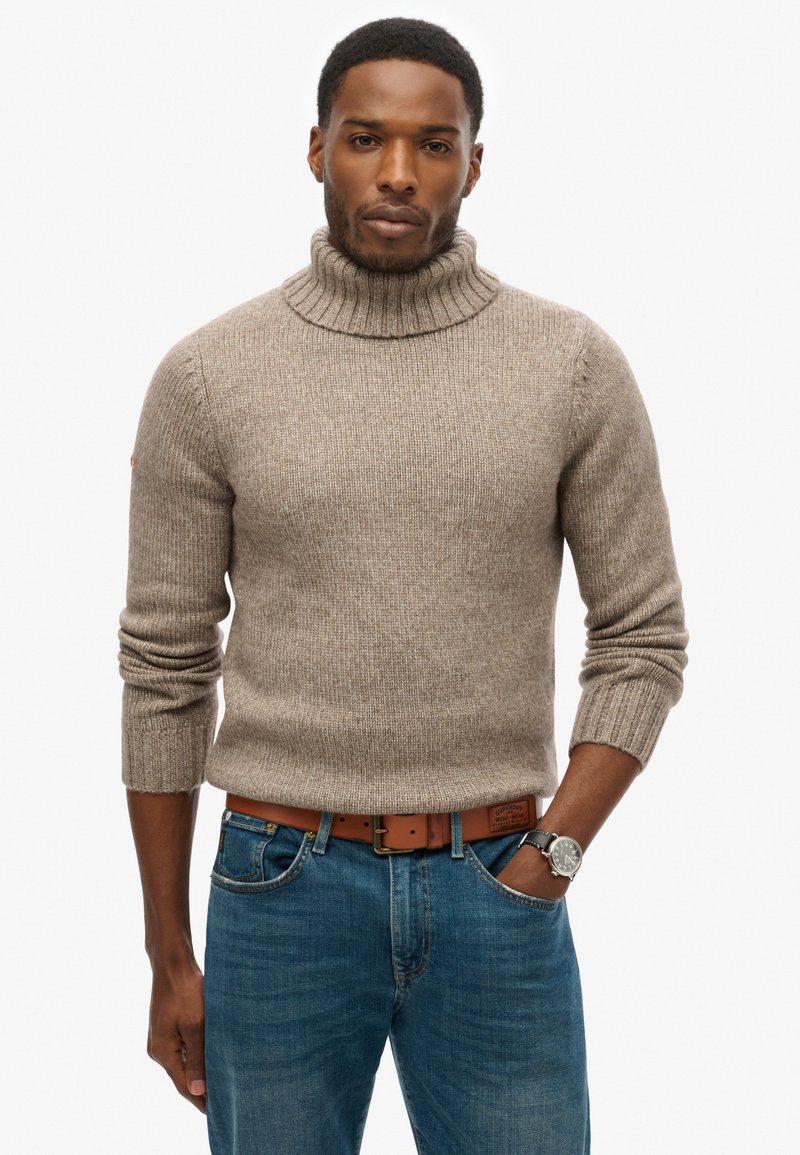 Suéter de punto beige con cuello alto, mangas largas y puños acanalados, combinado con jeans azules y un cinturón de cuero marrón.