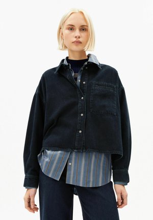 Junge Frau mit kurzen blonden Haaren, die eine dunkle, übergroße Jeansjacke trägt, darüber ein blau gestreiftes Hemd, vor weißem Hintergrund.