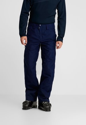 Homme portant un pantalon de ski bleu marine, des bottes de ski noires et un haut à manches longues foncé, debout devant un fond uni.