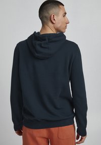 Tumši zils kapuces krekls, izgatavots no mīkstas auduma, ar auklu regulējamu kapuci un ribotu rokas piedurknēm. Aizmugurei ir vienkāršs, klasiskas formas dizains bez rotājumiem.