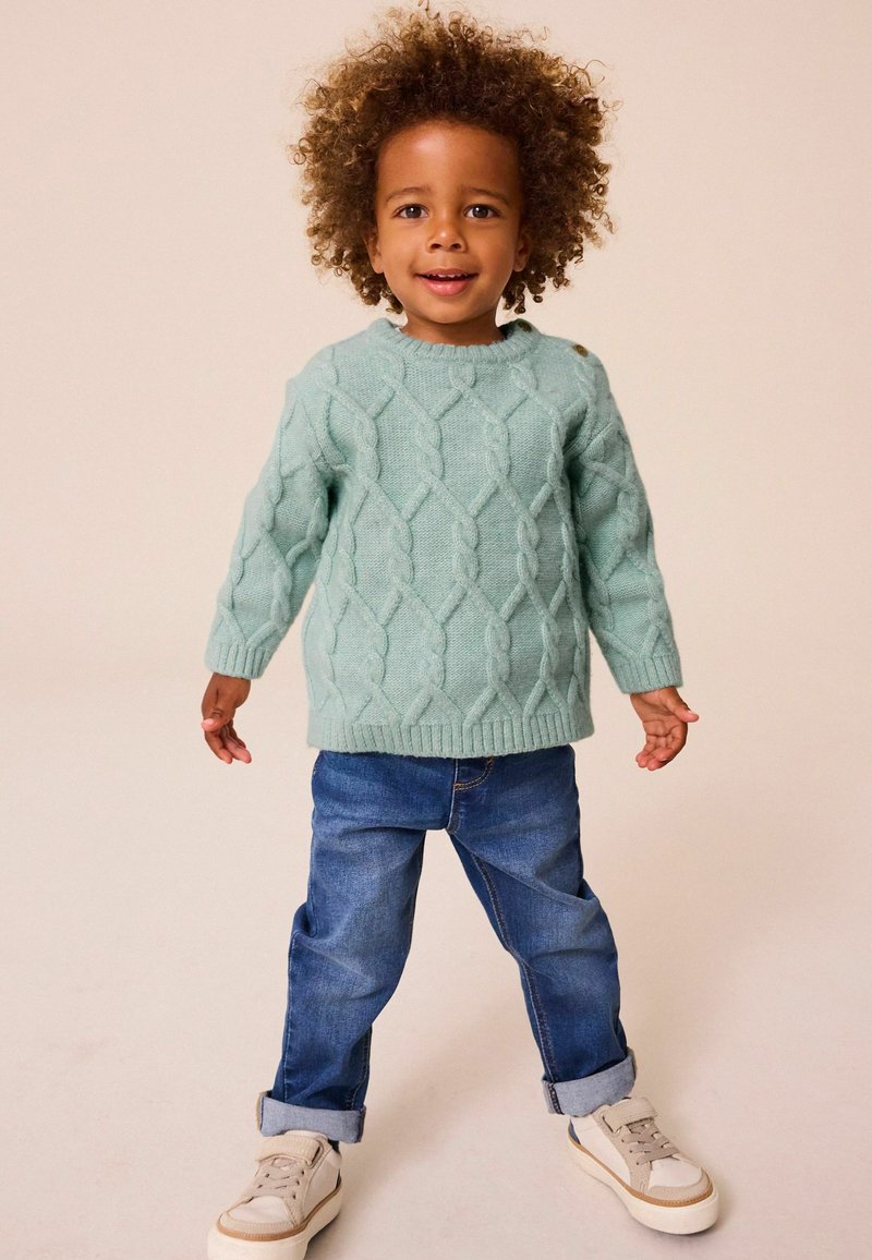 Bambino piccolo con capelli ricci che indossa un maglione a trecce verde chiaro, jeans blu risvoltati e scarpe da ginnastica beige, in piedi e sorridente.