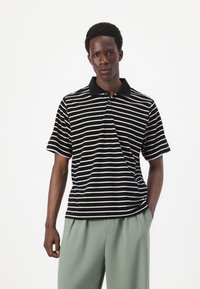 Black and white striped short-sleeve polo shirt with a black collar and three-button placket, paired with light green trousers.