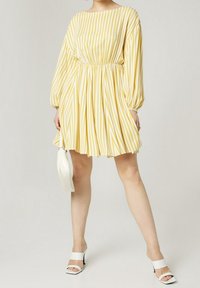 Žluté a bílé pruhované šaty s dlouhými rukávy a elastickým pasem. Spárované s bílými sandály na podpatku a malou bílou kabelkou.