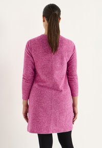 Rosa gestricktes Kleid mit langen Ärmeln und geradem Schnitt, ausgestattet mit einer Mittelnaht und einem gerippten Muster, kombiniert mit schwarzen Leggings.