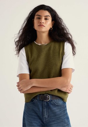 Donna con capelli lunghi e ricci che indossa orecchini a cerchio dorati, maglione senza maniche verde oliva sopra una T-shirt bianca e jeans blu con cintura nera.