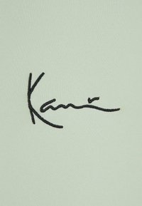 Un tissu vert clair présente une signature noire brodée avec des lignes fluides, mettant en avant une texture lisse et un design moderne.