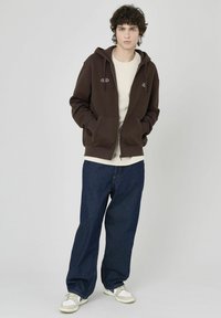 Haut à glissière marron avec cordon de serrage, poches avant et détail de logo, porté avec une chemise crème et un jean bleu ample. Baskets blanches.