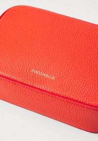 Červené kožené pouzdro s texturou a obdélníkovým tvarem. Má vyražené zlaté logo "Coccinelle". Šité okraje a hladký povrch.