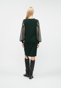 Σκούρο πράσινο φόρεμα με μακριά διάφανα μανίκια που διαθέτουν floral σχέδια, εφαρμοστή σιλουέτα και στρογγυλή λαιμόκοψη. Μαύρες μπότες μέχρι το γόνατο ολοκληρώνουν την εμφάνιση.