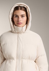 Puffová bunda svetlo béžovej farby s vysokým golierom, vypasovaným pásom a kapucňou. Vyznačuje sa hladkou textúrou a minimálnym šitím.