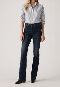 Femme portant un jean bootcut bleu foncé, une chemise à rayures à manches longues avec manches retroussées et des bottes noires à bout pointu.