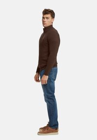Suéter de punto marrón con un alto cuello, combinado con jeans de mezclilla azules y zapatillas de suede marrón. Diseño sencillo, silueta ajustada.