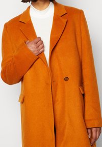 Abrigo de mezcla de lana naranja con cuello de muesca, cierre de dos botones y bolsillos laterales. Textura suave y silueta ajustada.