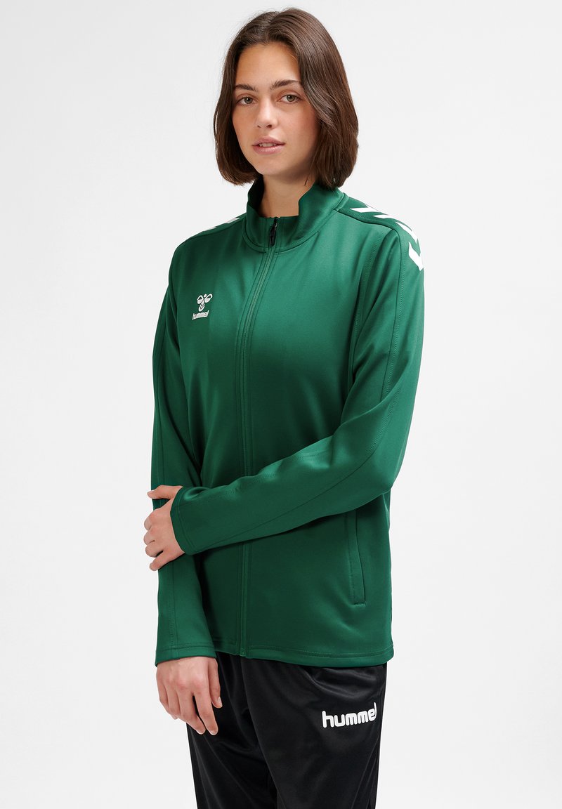 Green zip-up jacket with a high collar, made of smooth fabric. Features white side accents and logo on the left chest. Paired with black pants.