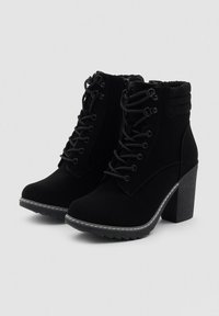 Botas de tobillo de ante negro con tacones gruesos, cuello acolchado, parte delantera con cordones, cremallera lateral y suela de goma texturizada con detalles de costura blanca.