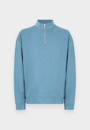Felpa azzurro chiaro con collo a mezza zip, maniche lunghe e polsini a costine. Presenta un logo discreto sul petto. Texture liscia.
