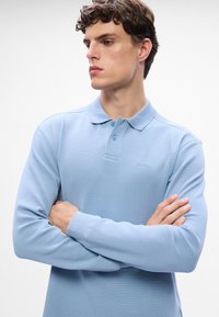 Gaiši zils garām piedurknēm polo krekls, izgatavots no tekstūru auduma, ar apkakli un divu pogu aizdari. Izšūts logotips uz krūtīm.